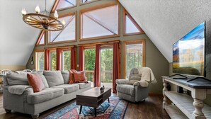 A spacious loft with a high ceiling, large windows, a cozy seating area with a sofa and armchairs, a wooden coffee table, and a television on a white stand. - Smoky Everest 4 Bedroom Chalet (Newport)