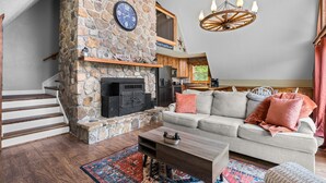 A cozy living room with a stone fireplace, a sofa with cushions, a coffee table, and a chandelier. - Smoky Everest 4 Bedroom Chalet (Newport)
