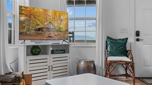 A living room with a television displaying a forest scene, a wicker chair, a white cabinet with a 'Believe' sign, and a view of the outdoors through a window.