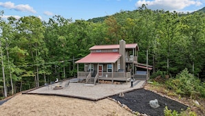 A two-story cabin with a red roof, a deck, and a fire pit area.