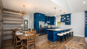 A modern kitchen with blue cabinets, a central island with stools, and a dining area with wooden furniture. - Deja Blue 2 3 Bedroom Home (Port Bolivar)