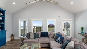 A spacious living room with a large sectional sofa, a glass coffee table, and a ceiling fan. There are three large windows providing views of the outdoors. - Deja Blue 2 3 Bedroom Home (Port Bolivar)