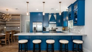 A modern kitchen with blue cabinets, a white island with stools, and pendant lighting. - Deja Blue 2 3 Bedroom Home (Port Bolivar)
