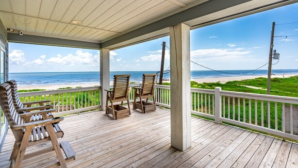 A wooden deck with chairs and a table, overlooking a beach with a clear sky. - Deja Blue 2 3 Bedroom Home (Port Bolivar)