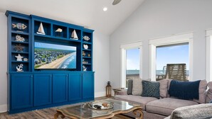 A living room with a large TV, blue shelving with decorative items, a glass coffee table, and a view of the beach through large windows. - Deja Blue 2 3 Bedroom Home (Port Bolivar)