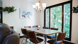 A dining area with a large table, brown leather chairs, a chandelier, and a view of trees through large windows.