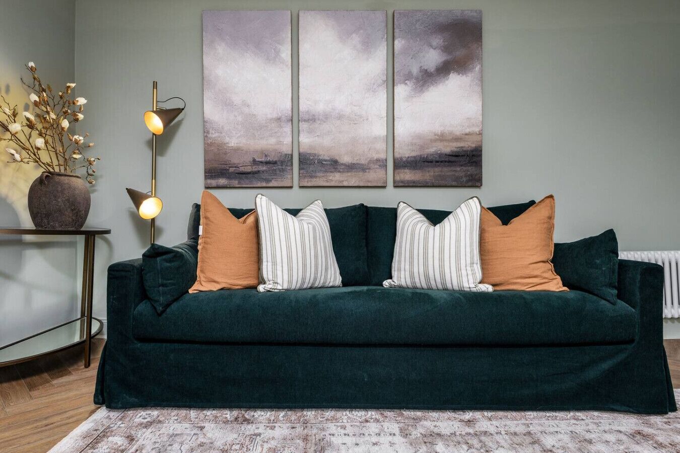 Un canapé vert foncé agrémenté de coussins à rayures et unis, un lampadaire et un vase avec des branches, le tout posé sur une table d’appoint.
