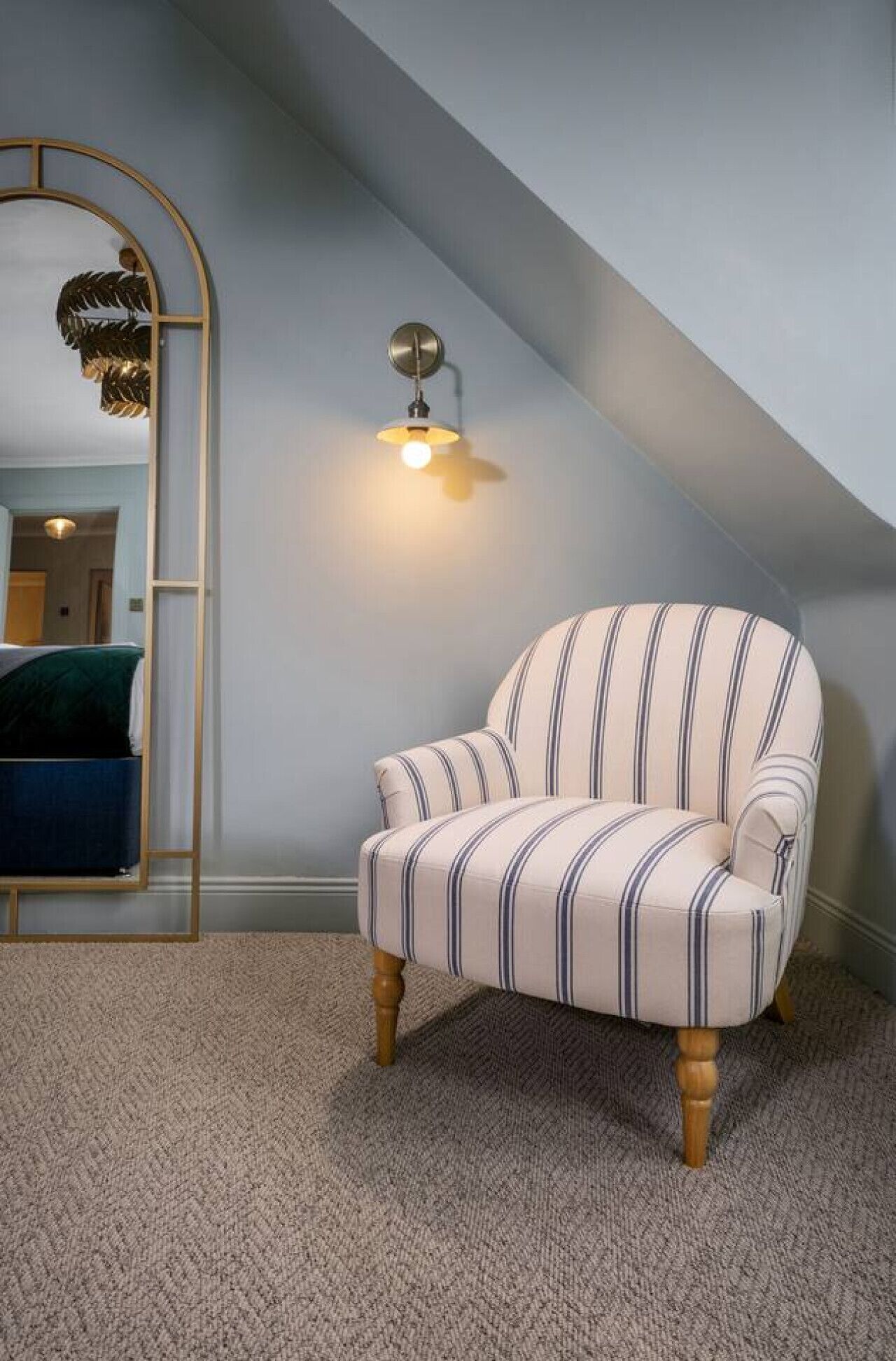 A corner room with a striped armchair, a wall-mounted light fixture, and a mirror reflecting a bed and a door.