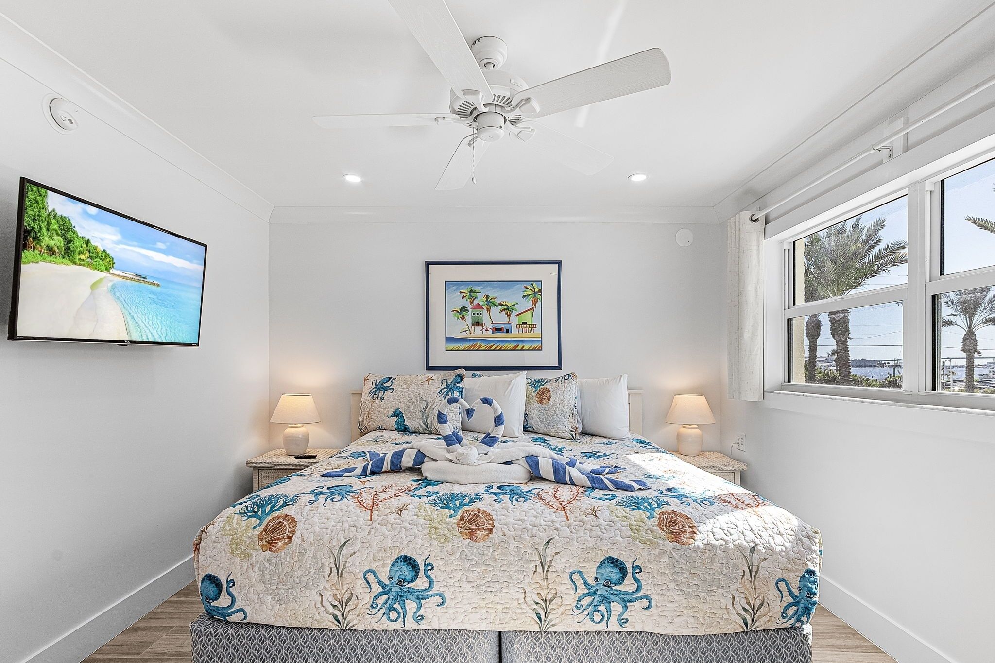 A bedroom with a bed, ceiling fan, wall-mounted TV, framed artwork, bedside lamps, and a window with a view.
