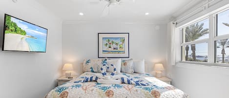 A bedroom with a bed, ceiling fan, wall-mounted TV, framed artwork, bedside lamps, and a window with a view.