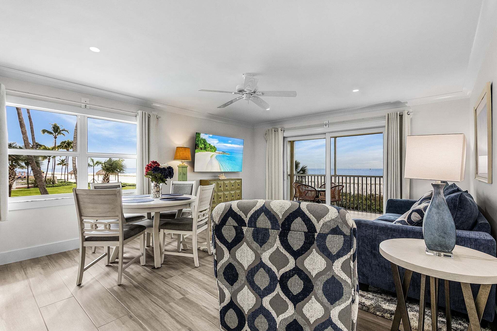 A modern living room with a dining area, large windows with a view of palm trees and the ocean, a ceiling fan, and a TV mounted on the wall.