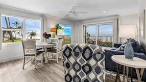 A modern living room with a dining area, large windows with a view of palm trees and the ocean, a ceiling fan, and a TV mounted on the wall. - Vacation Villas #131 2 Bedroom Condo (Fort Myers Beach)