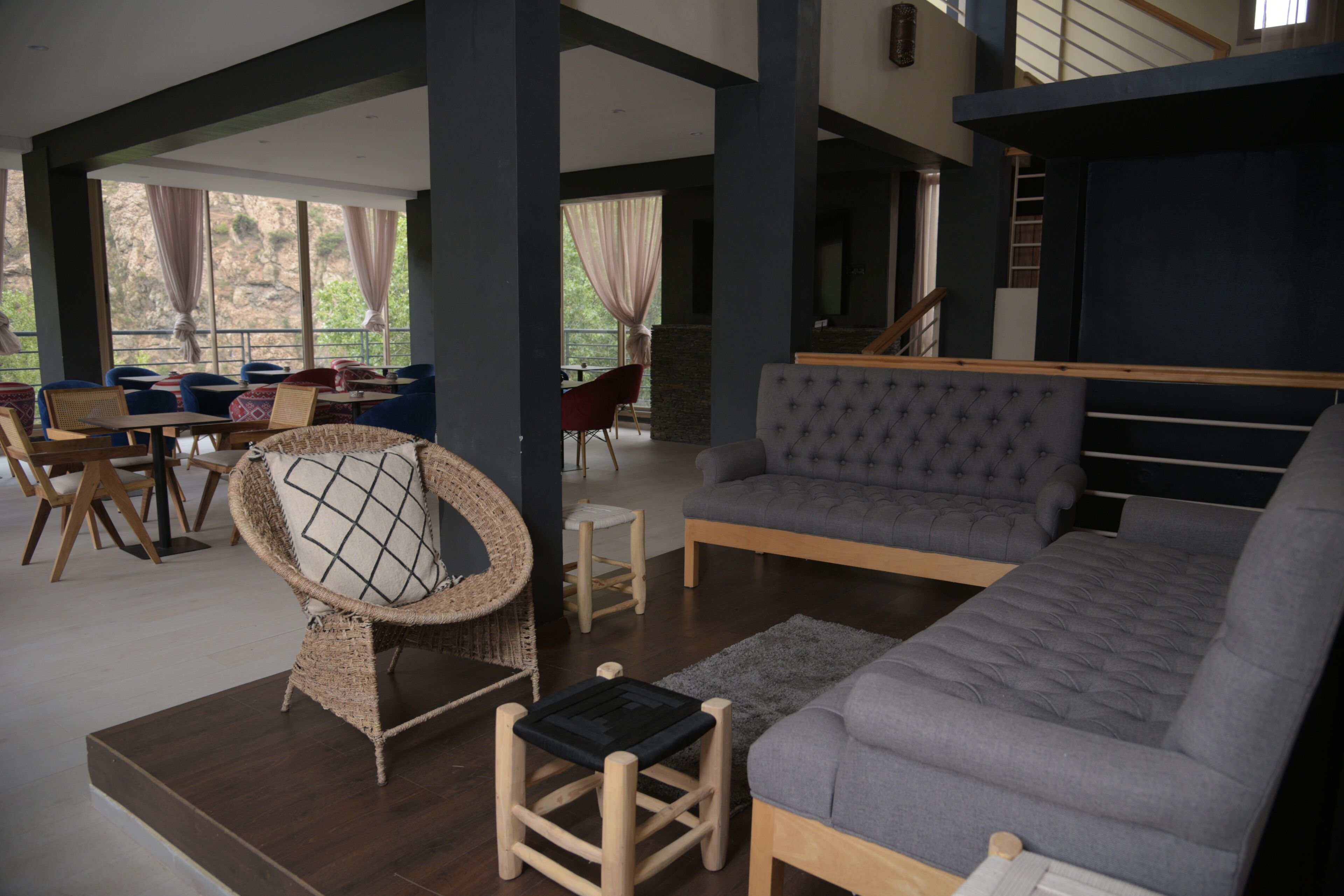 Amplia sala de estar con un sofá gris, un sillón de mimbre con cojín y una pequeña mesa de madera. Se ve una escalera al fondo que conduce a un nivel superior.