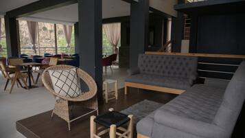 Amplia sala de estar con un sofá gris, un sillón de mimbre con cojín y una pequeña mesa de madera. Se ve una escalera al fondo que conduce a un nivel superior.