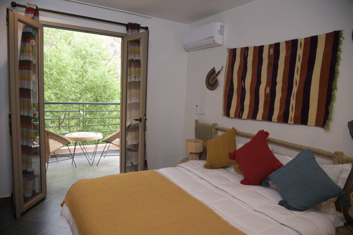 A bedroom with a bed, pillows, a wall hanging, and a balcony with chairs.