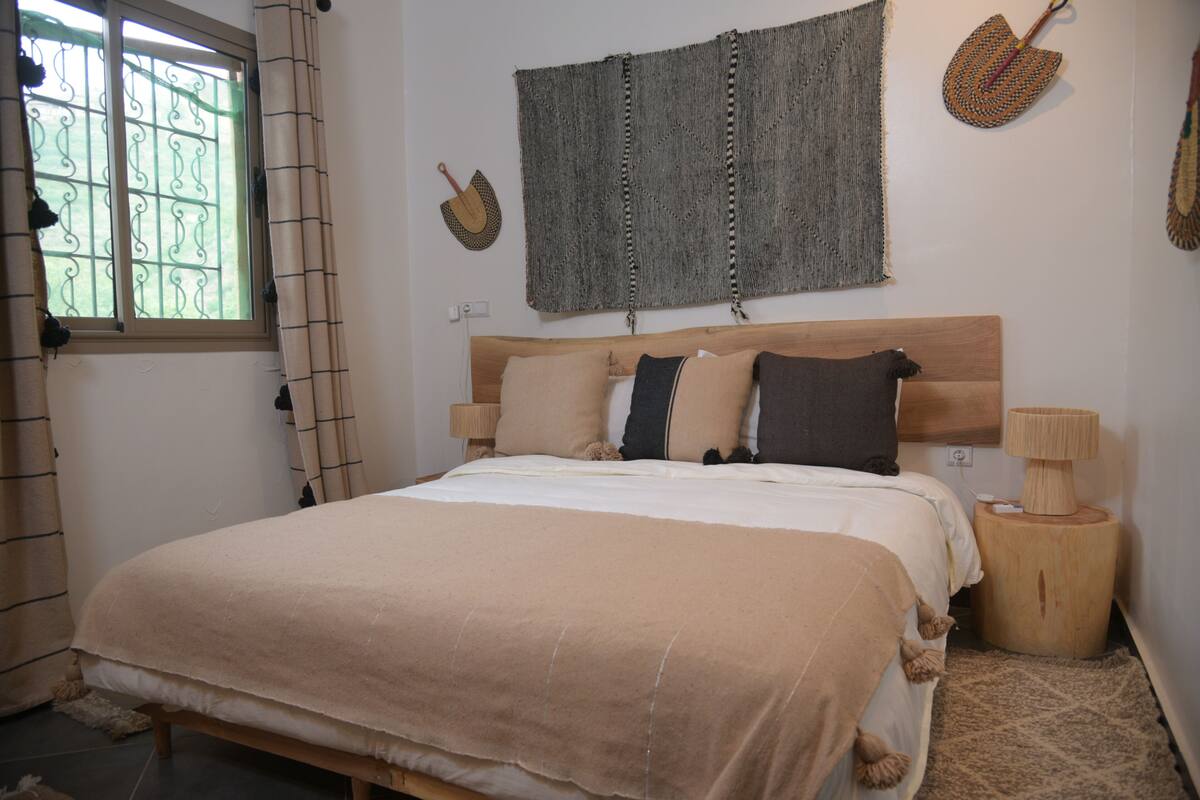 A bedroom with a bed, a headboard, a bedside table, a lamp, a wall hanging, and a window with curtains.