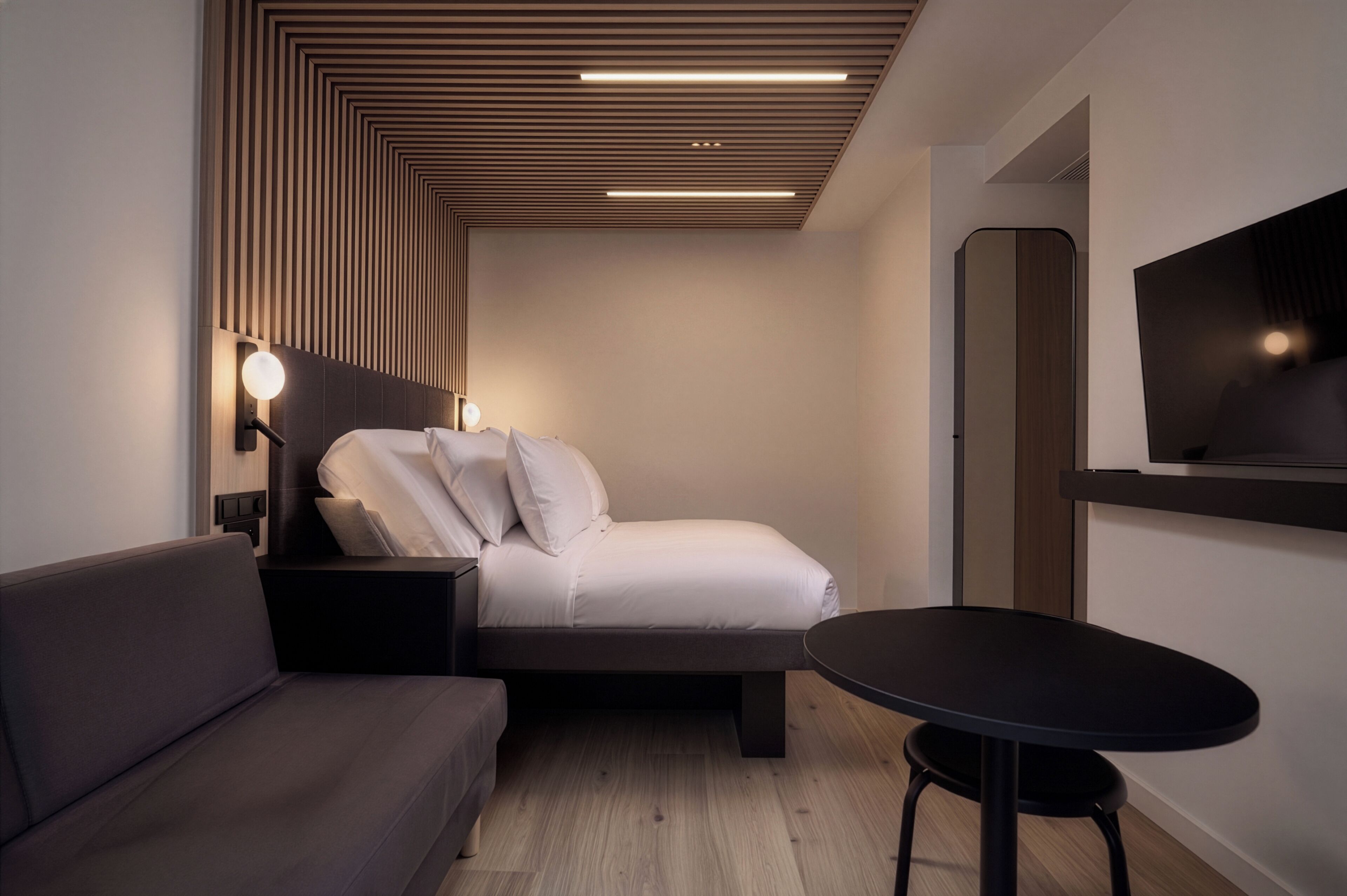A modern hotel room with a bed, a sofa, a round table, and a wall-mounted TV.