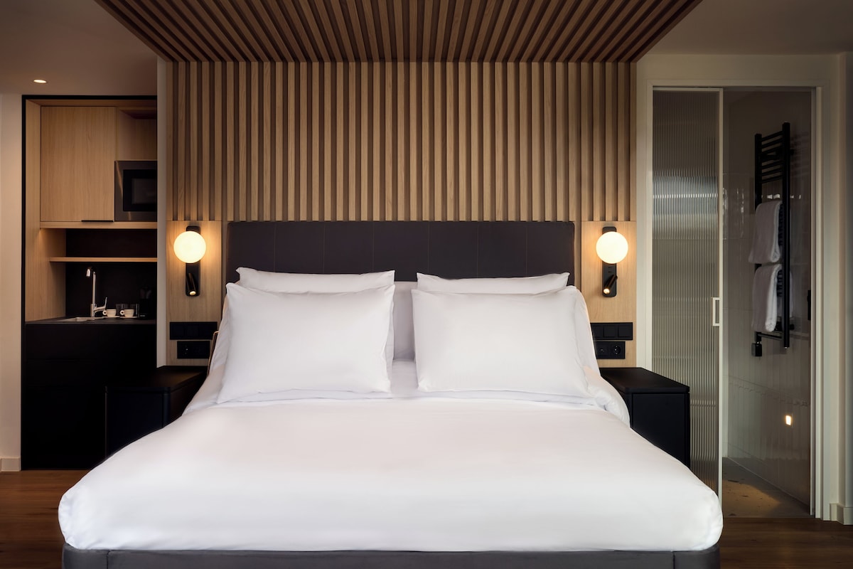 A modern hotel room with a large bed, bedside lamps, a built-in TV, and a shower area.