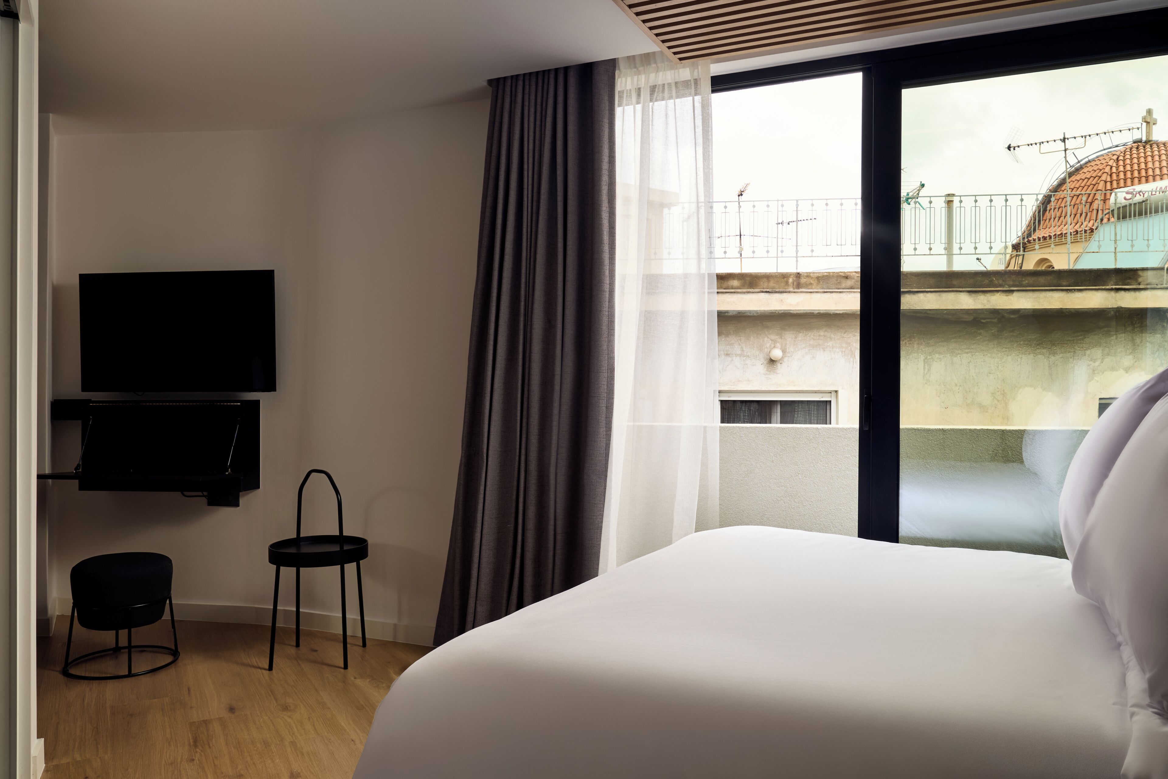 A hotel room with a bed, a TV mounted on the wall, a small stool, and a view of a balcony with a railing.