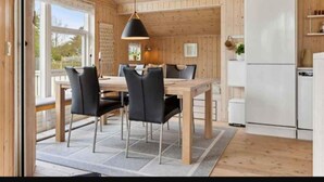 A modern kitchen with wooden walls and a large window, a dining area with a table and chairs, and a white refrigerator.