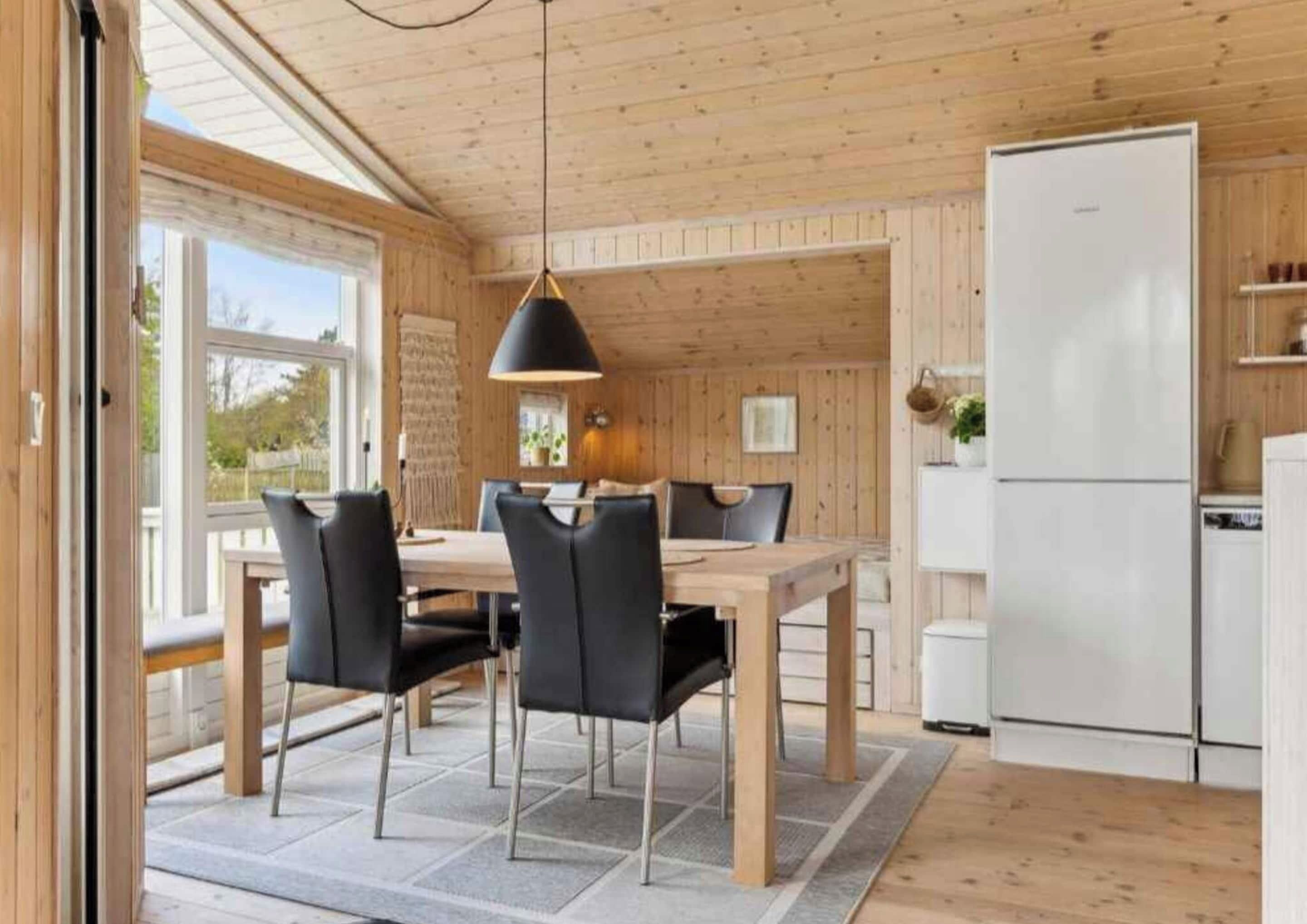 A modern kitchen with wooden walls and a large window, a dining area with a table and chairs, and a white refrigerator.