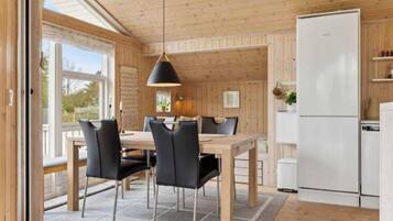 A modern kitchen with wooden walls and a large window, a dining area with a table and chairs, and a white refrigerator.