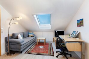 Habitació compacta amb sostre inclinat, un lucernari, un sofà, escriptori, cadira, làmpada de peu i taula petita.