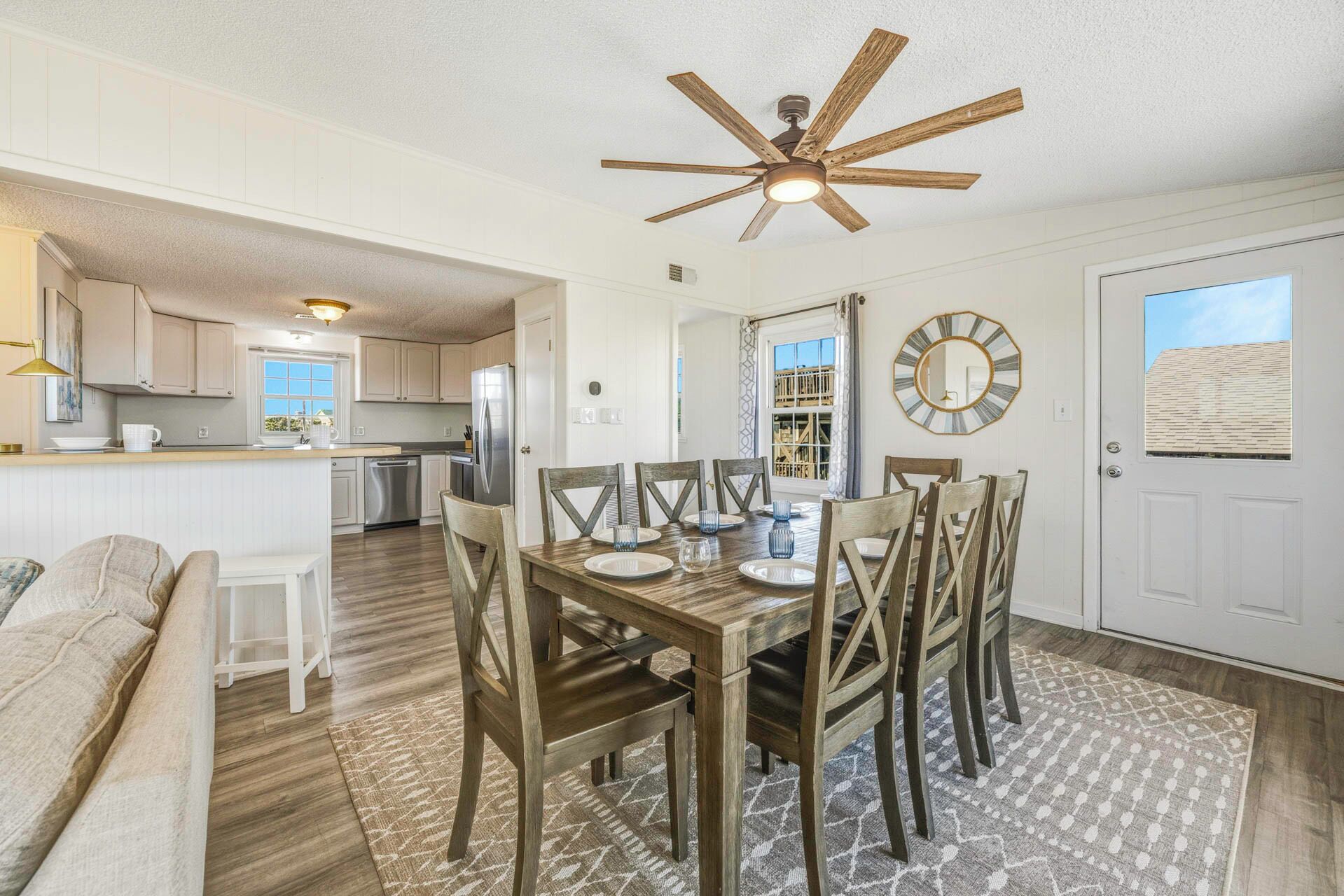 A dining area with a wooden table, chairs, and a ceiling fan. There is a kitchen in the background with white cabinets and a refrigerator.