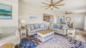 A spacious living room with a large sofa, a coffee table, and a patterned rug. There is a dining area with a table and chairs, and a kitchen visible in the background. - Oceanfront Home in Avon with Hot Tub and Stunning Views (Avon)