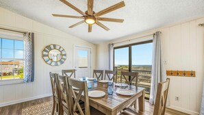 A dining area with a wooden table and chairs, a ceiling fan, and a view through a large window. - Oceanfront Home in Avon with Hot Tub and Stunning Views (Avon)