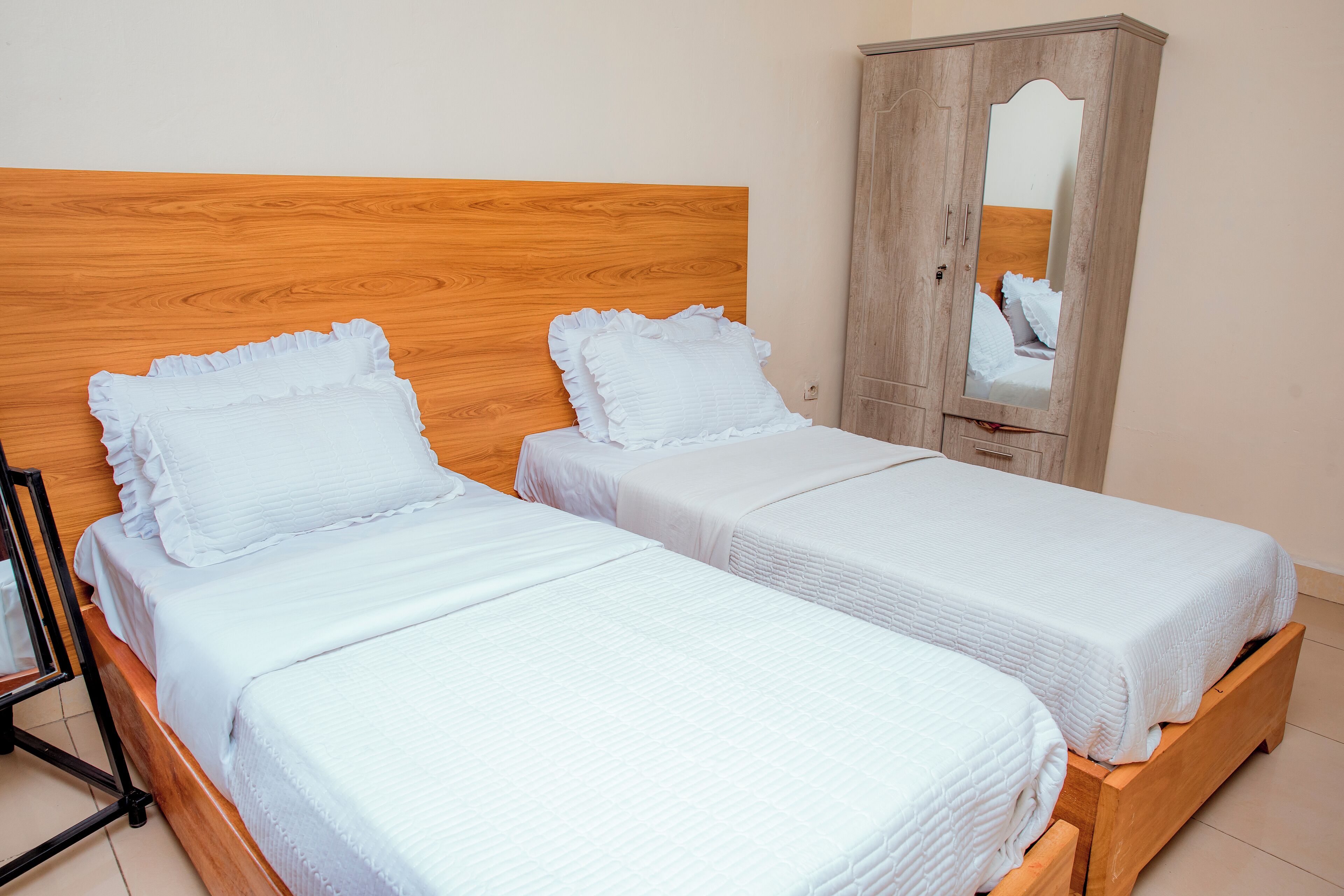 Two single beds with wooden headboards and white bedding, a wooden wardrobe with a mirror, and a black metal frame to the left.