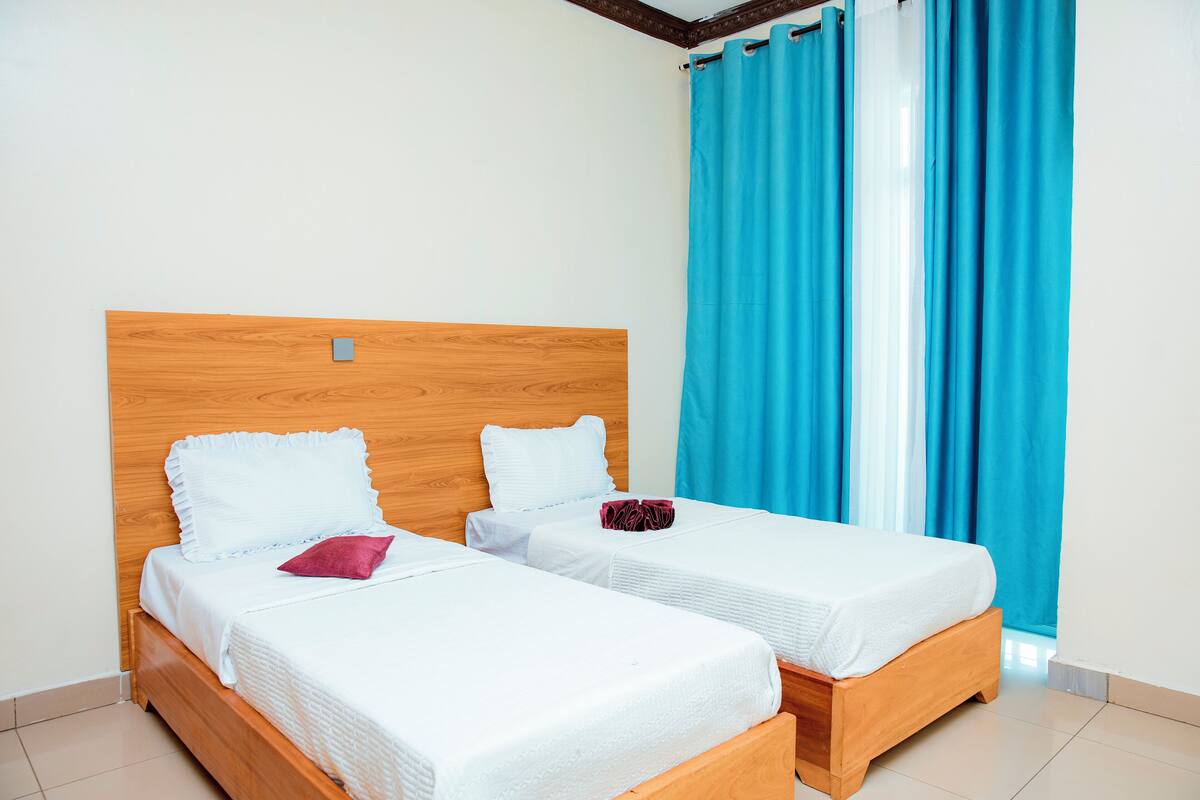 A room with two single beds, wooden headboards, white bedding, and blue curtains.