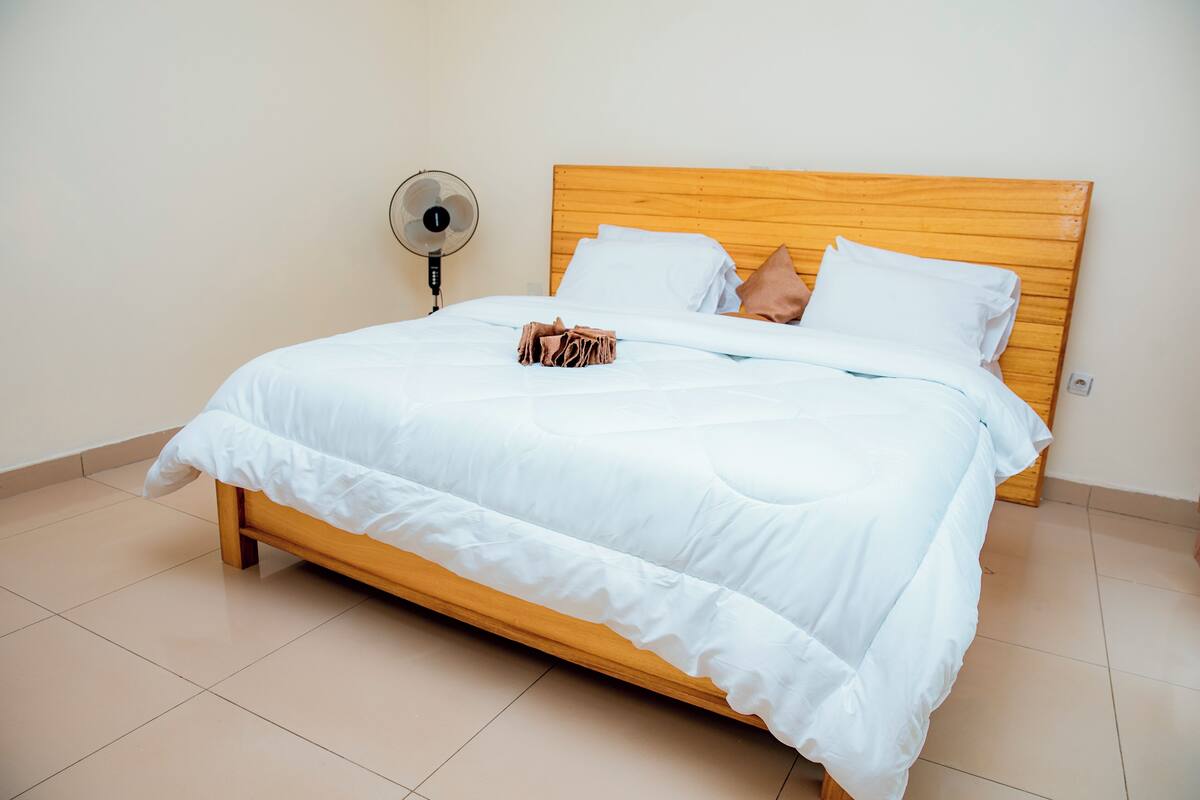 A neatly made bed with white bedding, a wooden headboard, and a fan on the wall.