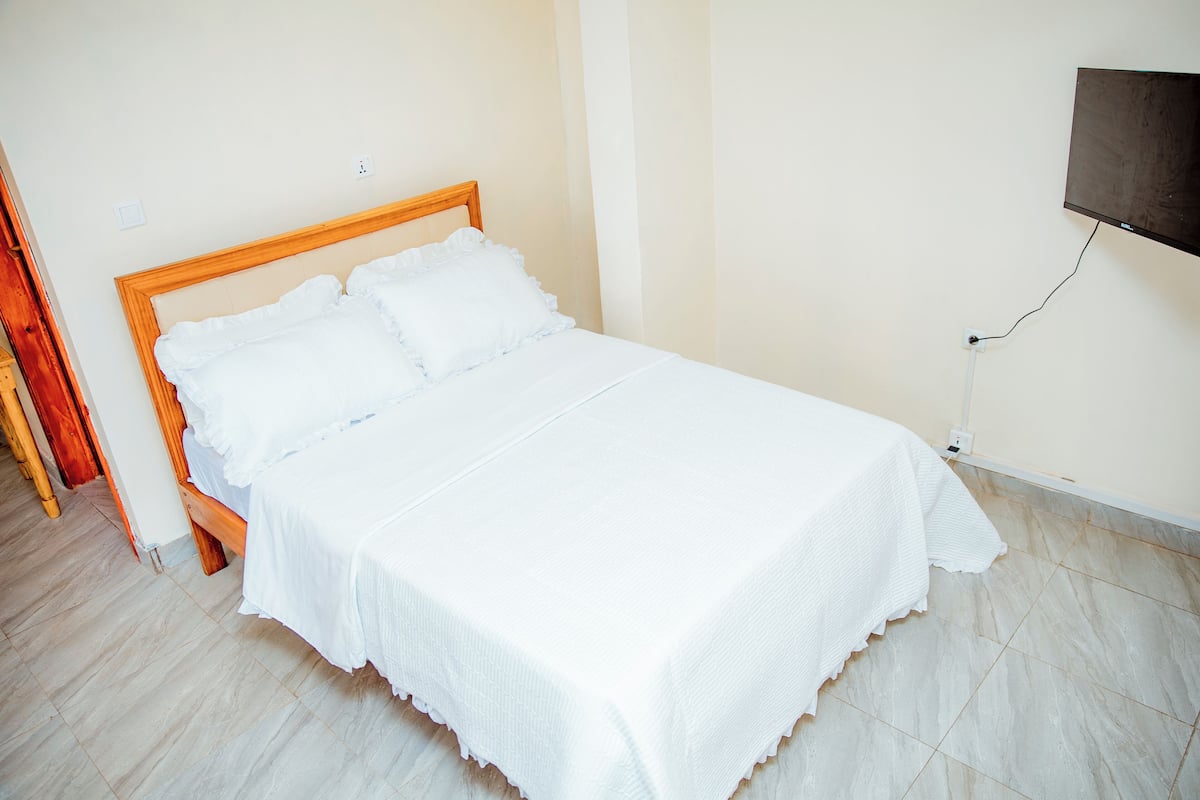 A neatly made bed with a wooden headboard, a flat-screen TV mounted on the wall, and a tiled floor.