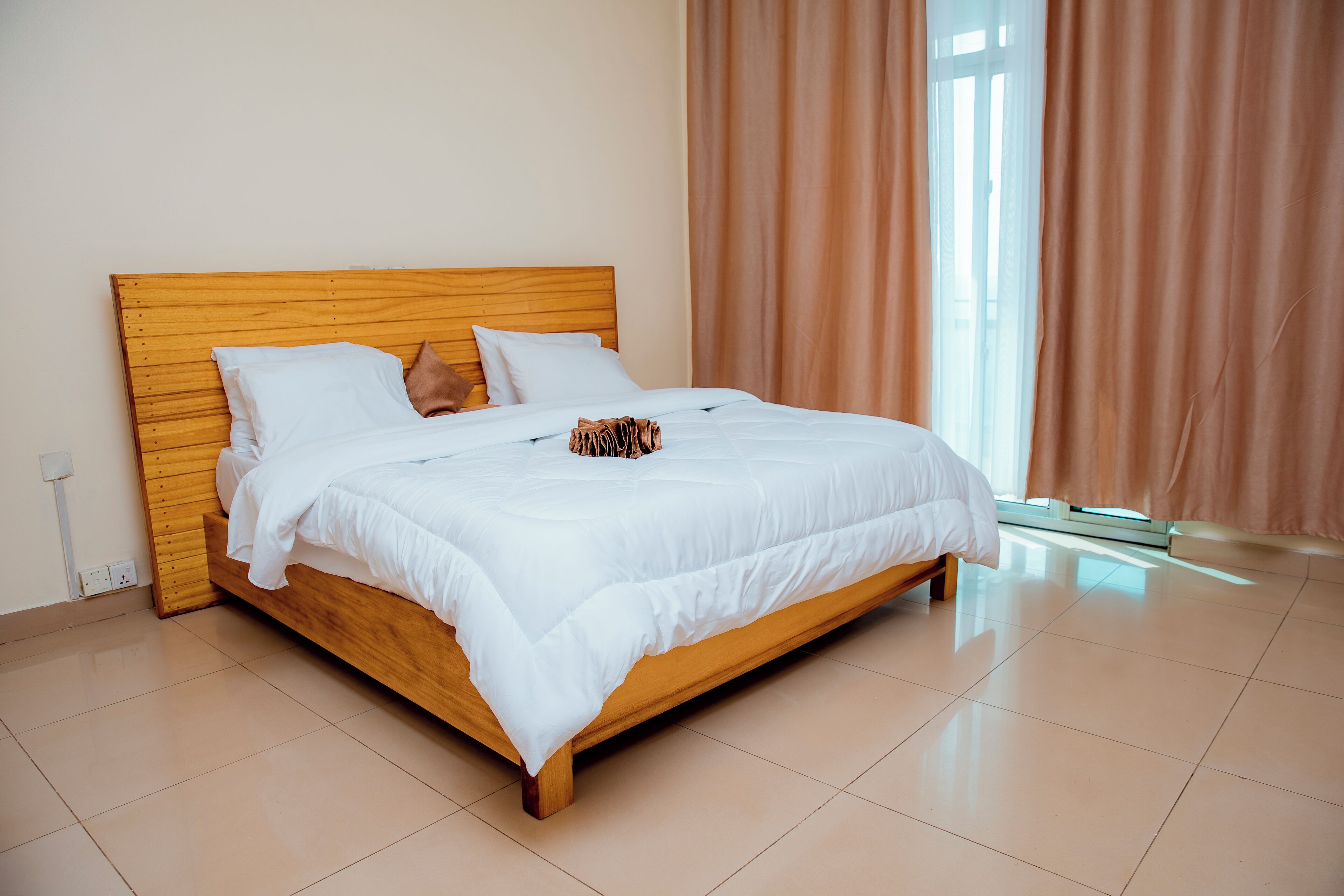 A neatly made bed with white bedding and a wooden headboard in a bright room with a large window and curtains.