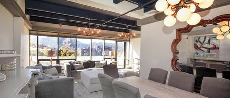 A modern dining area with a large table and chairs, a chandelier, and a view of the cityscape through large windows.