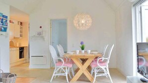 Et spiseområde med et træbord og hvide stole, en hængende lampe samt udsigt til et køkken og en opholdsstue.