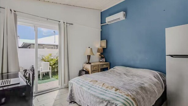 A hotel room with a bed, a desk, a chair, a nightstand, a lamp, and an air conditioning unit. - Al Capone - Paradise Apartment (Cape Coral)