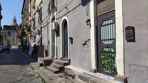 A narrow street with buildings on either side, a blue door with a barred window, and a small balcony with a blue towel.