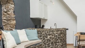 A cozy living area with a stone wall, a sofa with cushions, a white cabinet, and a kitchen area with a sink and countertop.