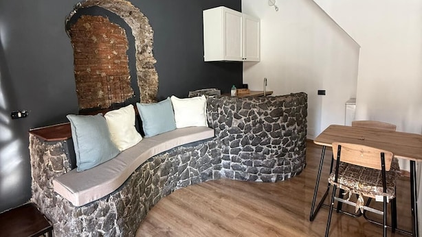 A cozy, stone-walled seating area with a built-in bench and cushions, a wooden dining table with chairs, and a white cabinet above. - Jesuit Home - Lumina Collection (Catania)