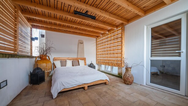 Una camera da letto con soffitto in legno, un letto con biancheria bianca, un vaso grande e una porta scorrevole in vetro.