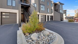 A modern multi-story residential building with a garage entrance, a staircase, and a landscaped area with rocks and shrubs. - Newly Constructed Condo along the Blue River (Silverthorne)