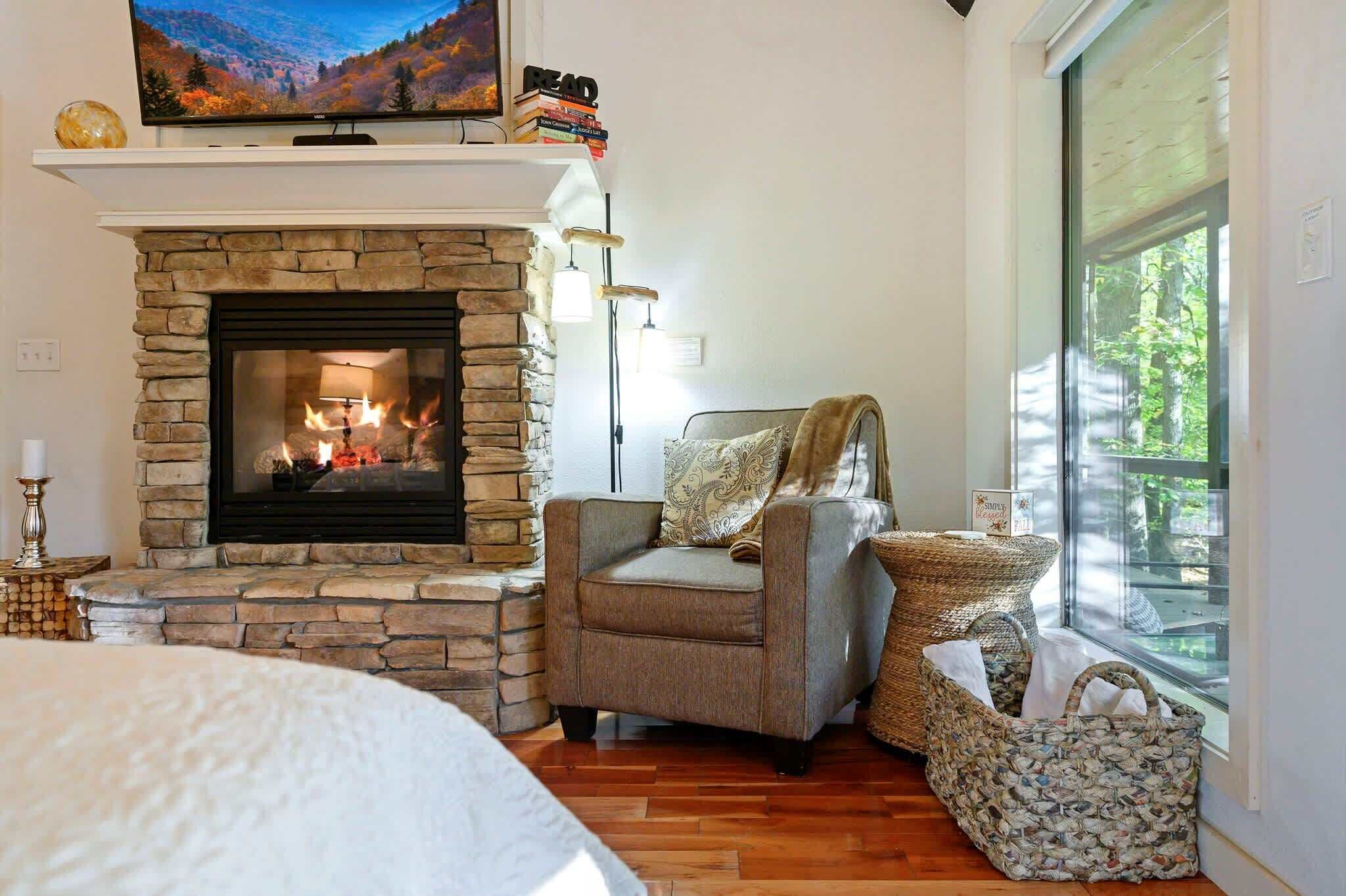 A cozy room with a stone fireplace, a comfortable armchair, a large window with a view of greenery, and a television on a shelf above the fireplace.