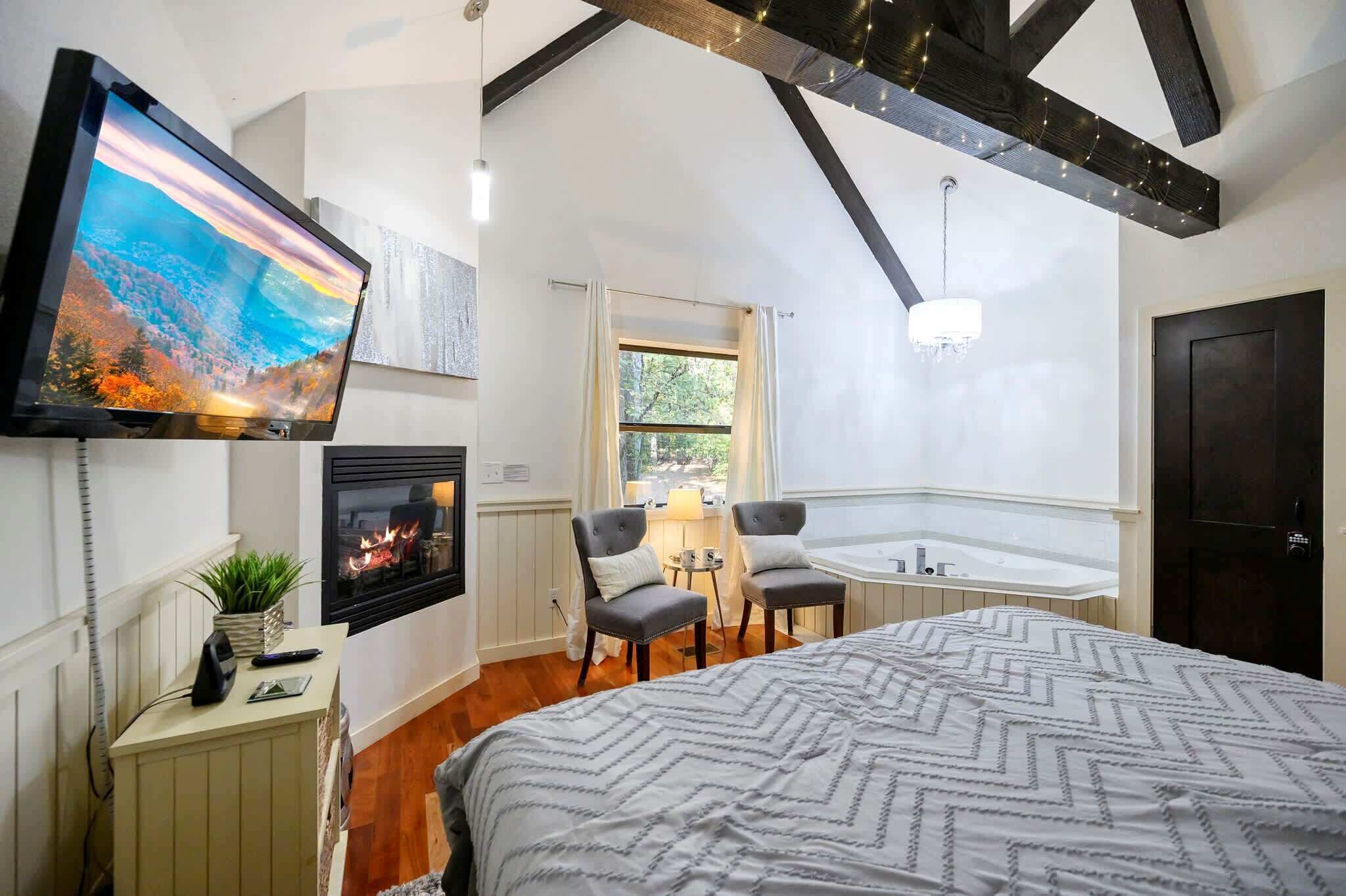 A bedroom with a large bed, a fireplace, a TV, and a small seating area with a view of the outdoors.