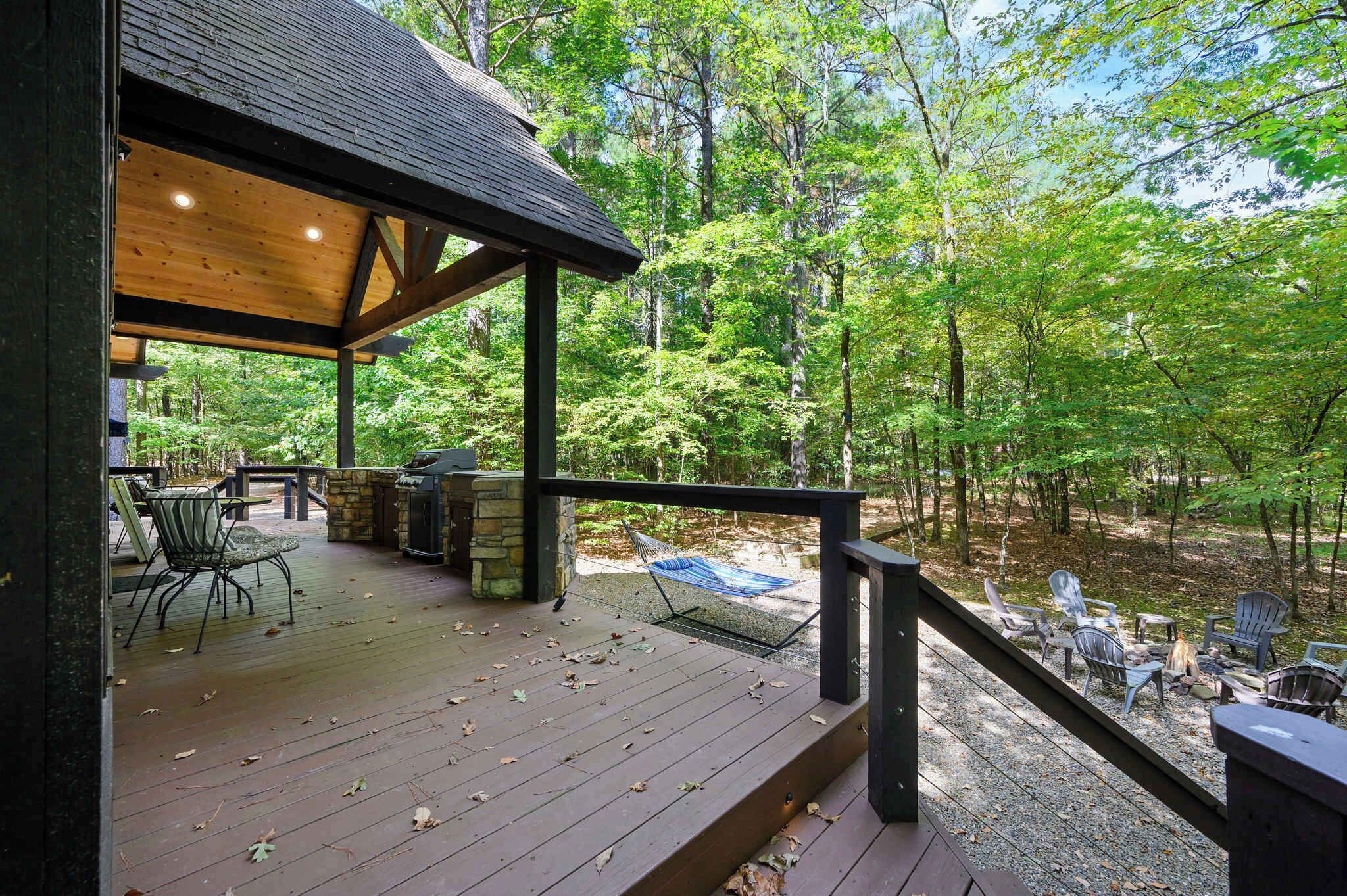A wooden deck with outdoor furniture, a stone fireplace, and a view of a forest.