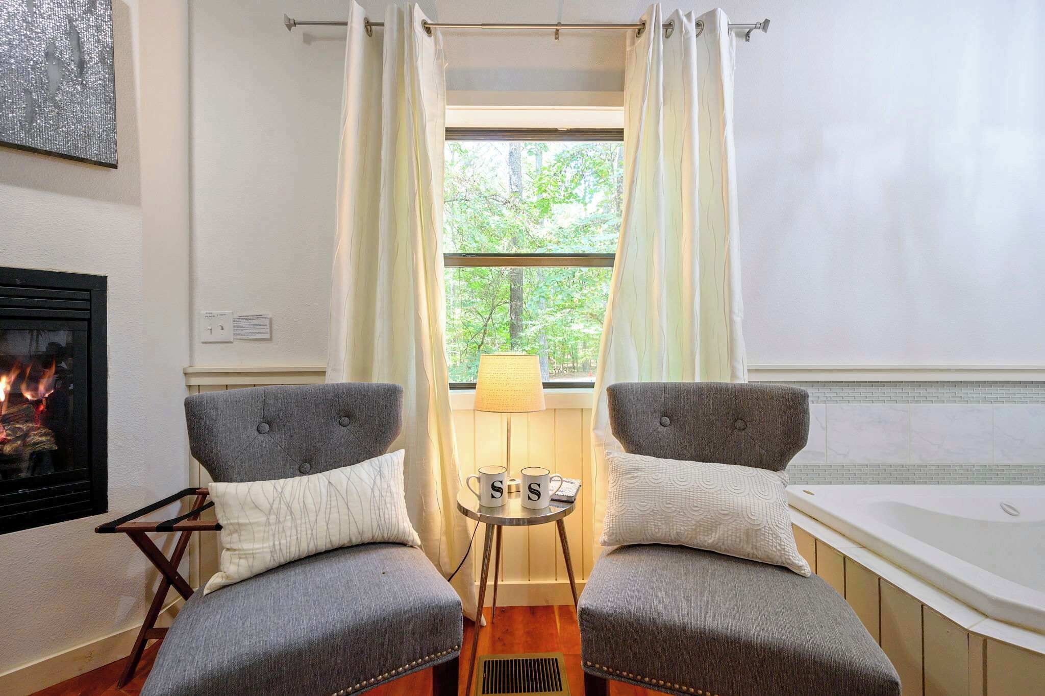A cozy room with two grey armchairs, a small table with a lamp, a fireplace, and a window with curtains.
