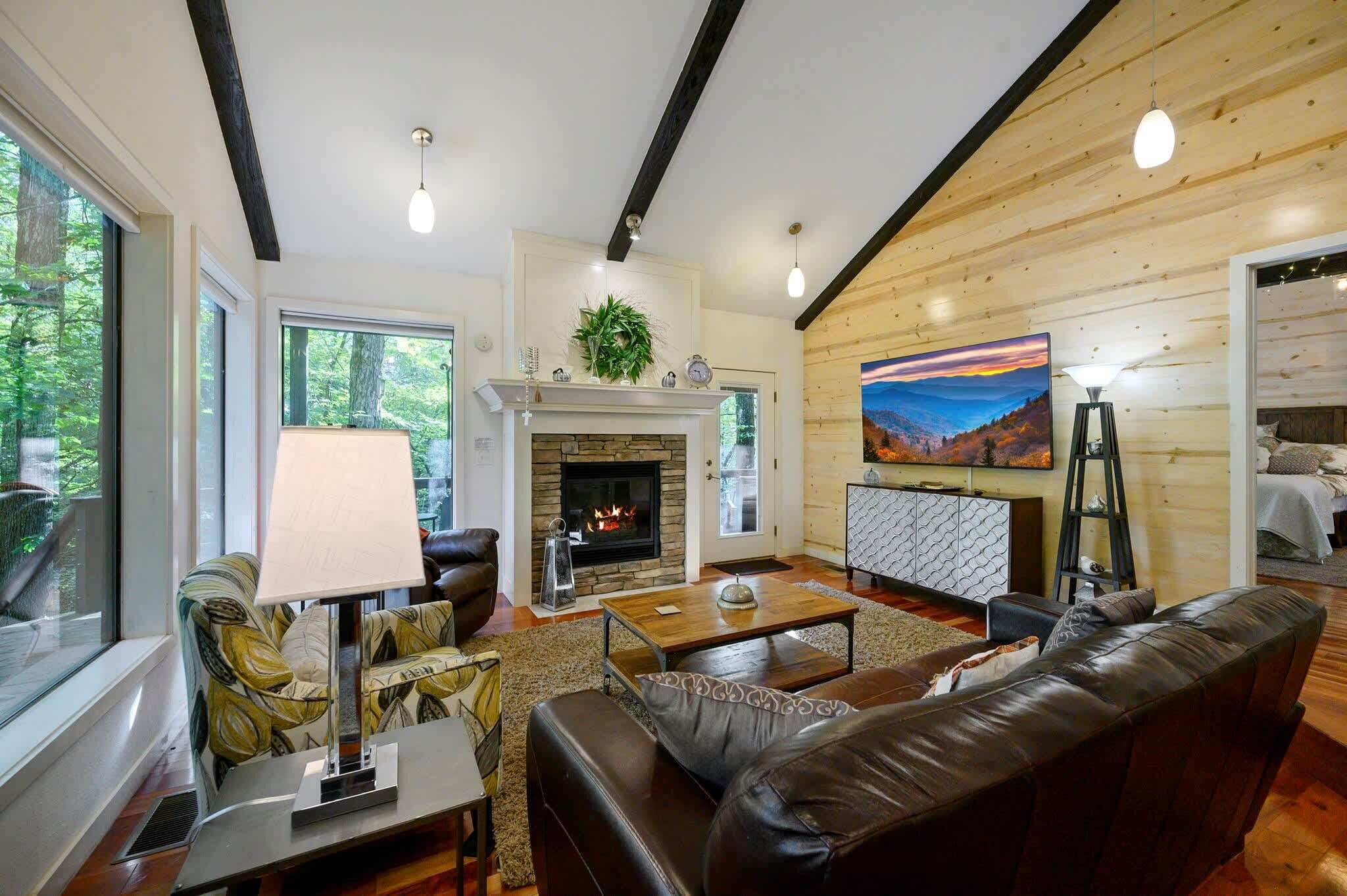 A cozy living room with a fireplace, leather sofa, and a large window overlooking greenery.