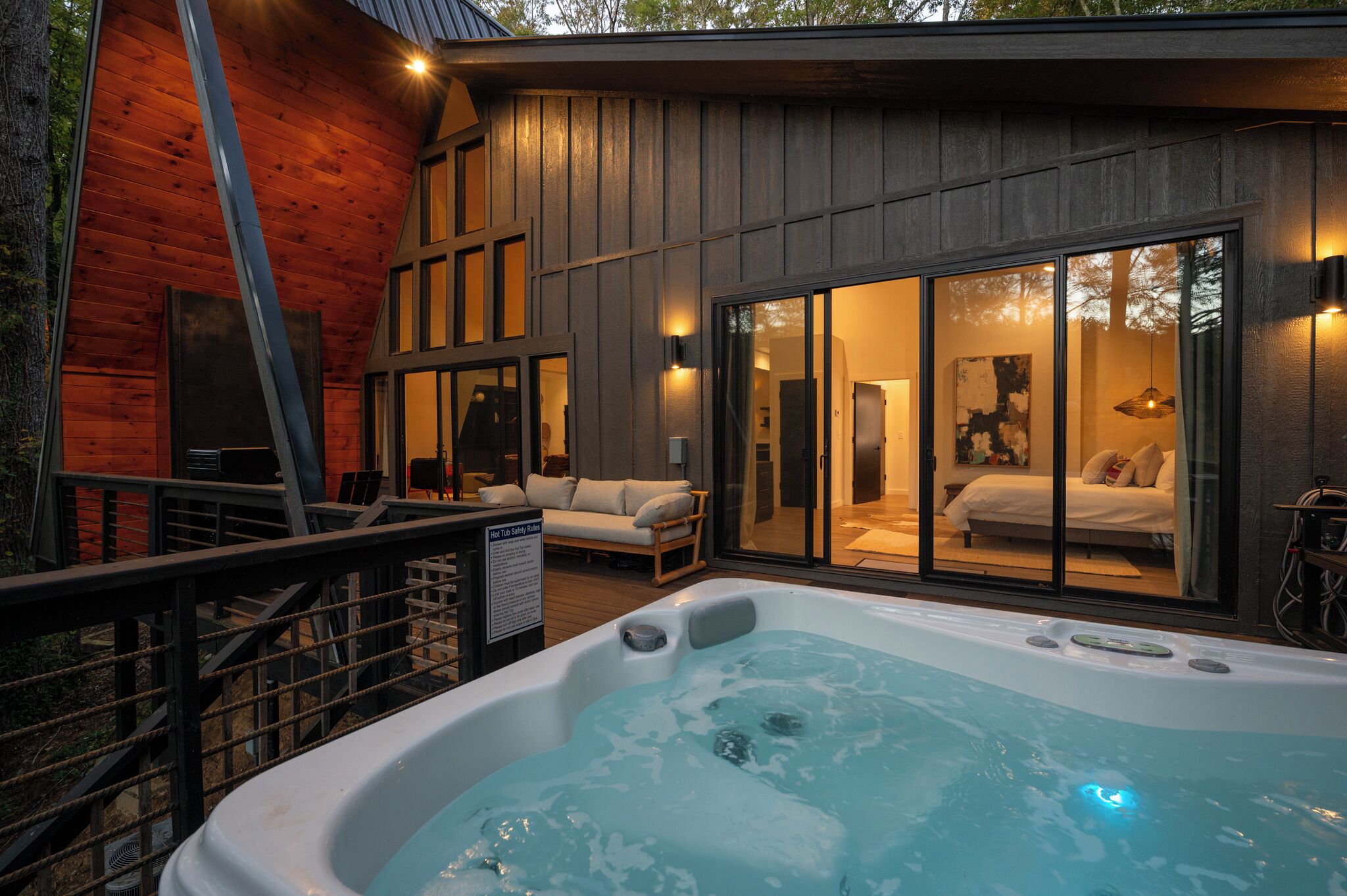 A modern cabin with a hot tub, wooden exterior, and large sliding glass doors leading to a bedroom with a bed and a bathroom.