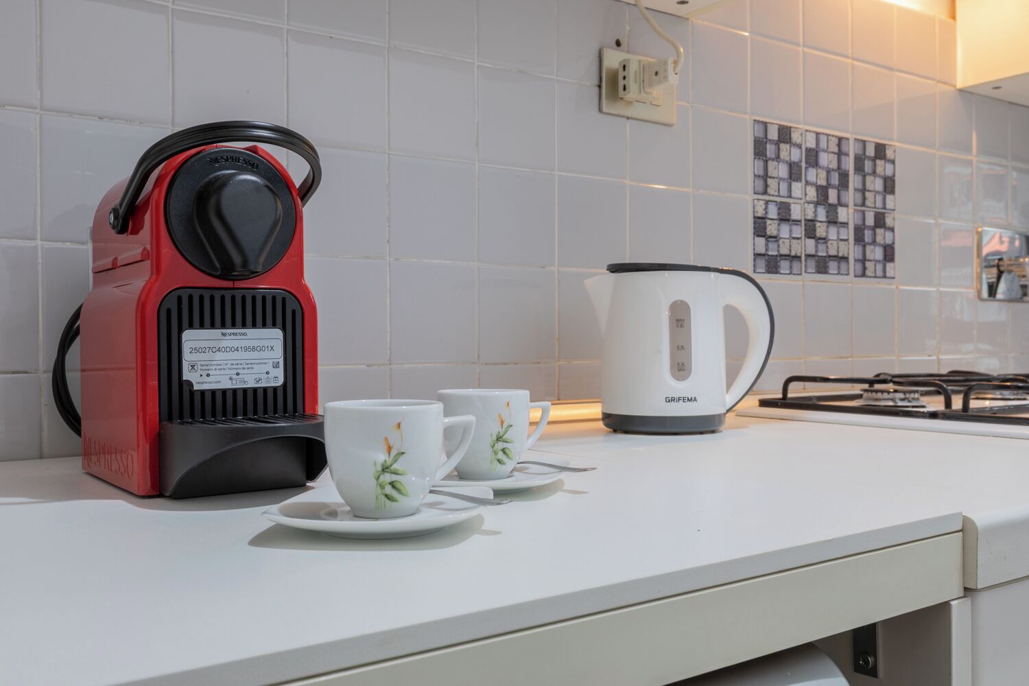 A kitchen with a red espresso machine, a white electric kettle, and two white cups with floral designs on a white countertop.